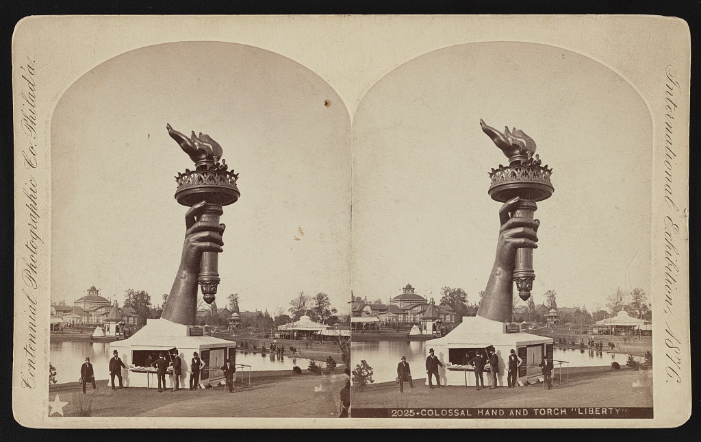 stereograph of large arm carrying torch next to a small building on fairgrounds