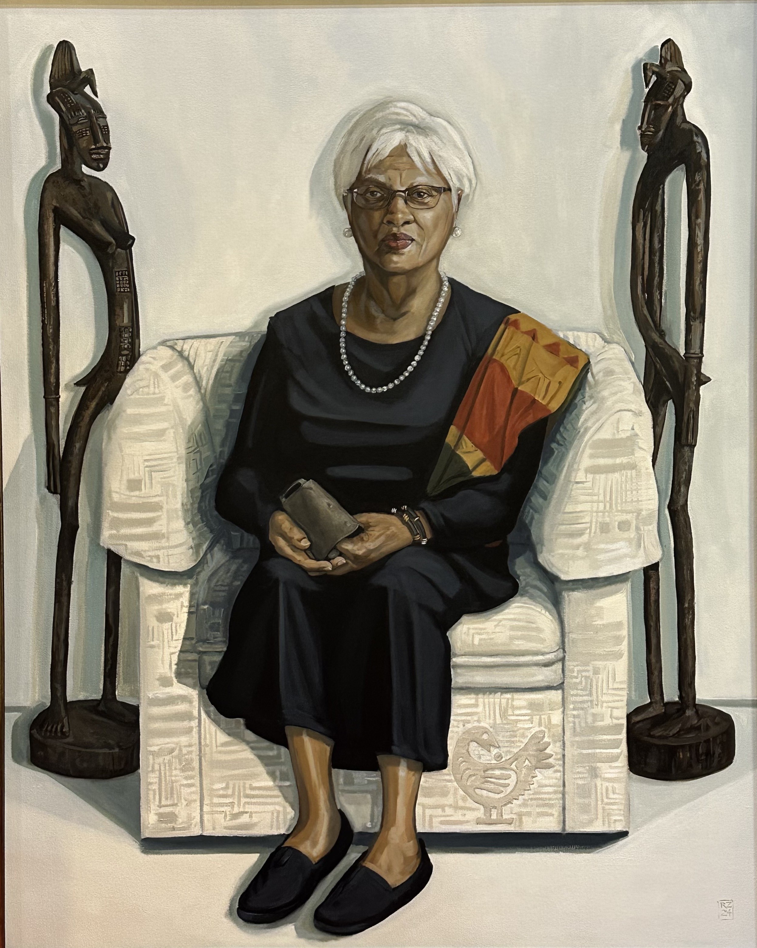 Oil painting of a woman sitting on a white chair holding a cowbell and flanked by two African figural sculptures.