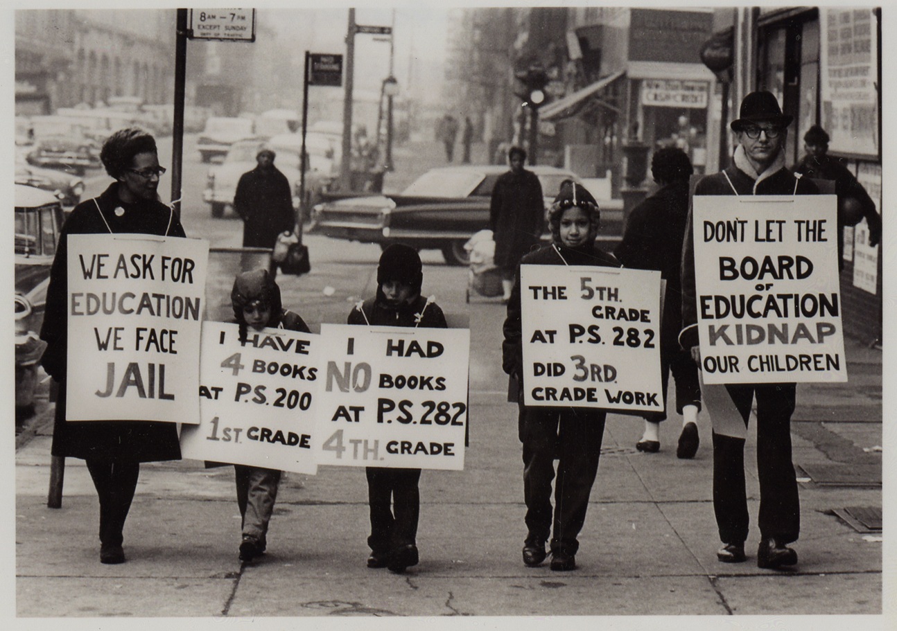 Black and white photograph of a woman, a man and their three children wearing placards describing education injustice while waking down a Brooklyn sidewalk.