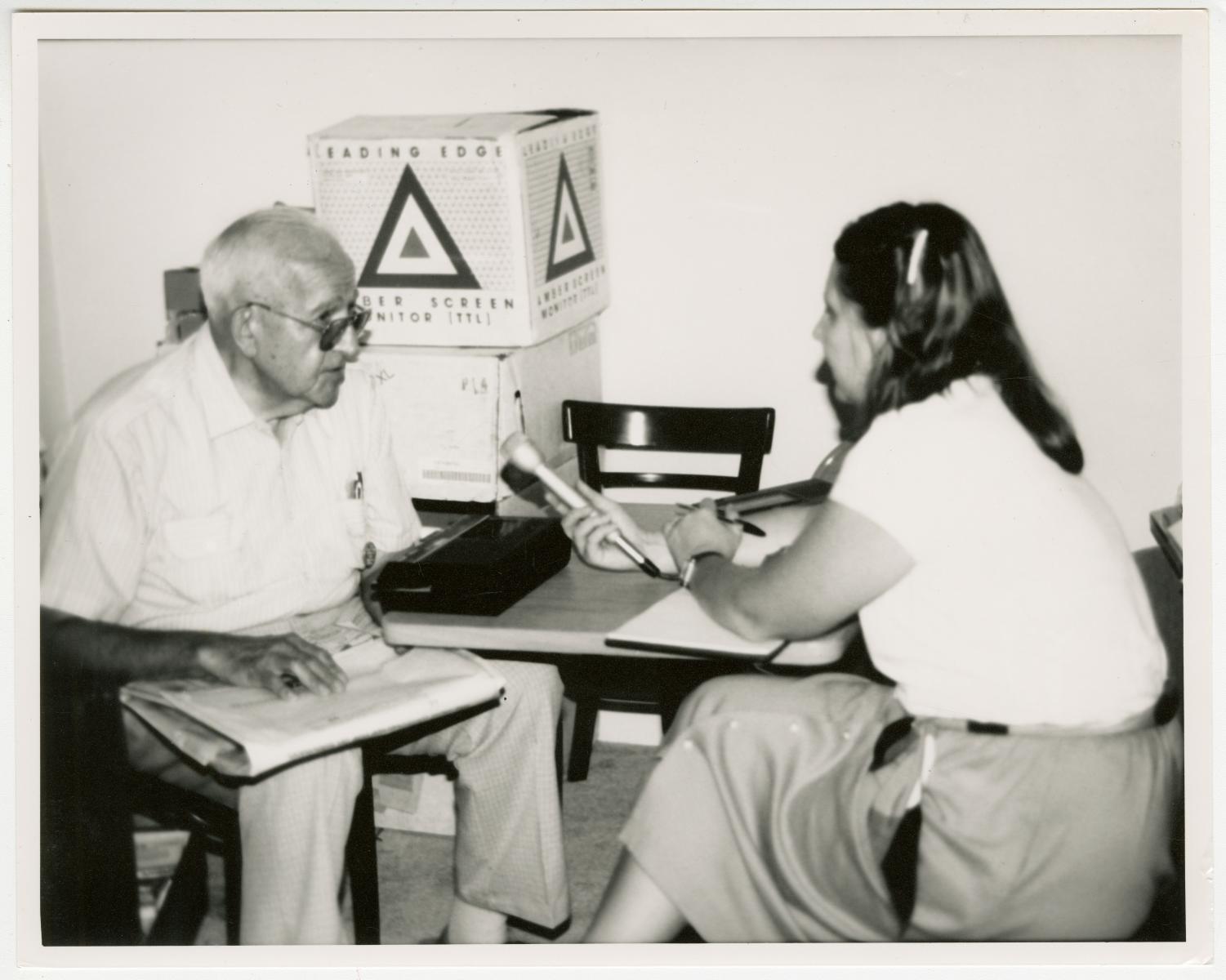 Black and white photograph of a woman holding a microphone and recording an older man as he's speaking.