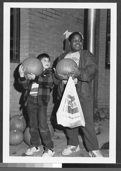 two boys, one white, one African American each holding a pumpkin near a brick wall