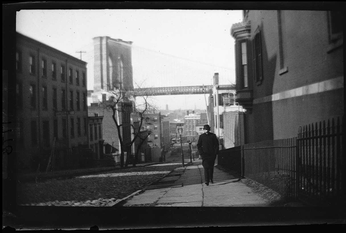Black and white photograph of a Brooklyn Heights cobblestone street looking towards the Brooklyn Bridge in the background, with a man walking up the sidewalk in the midground.