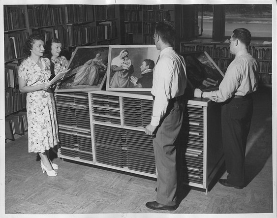 Black and white photograph of people examining framed reproductions of artwork amidst library bookshelves