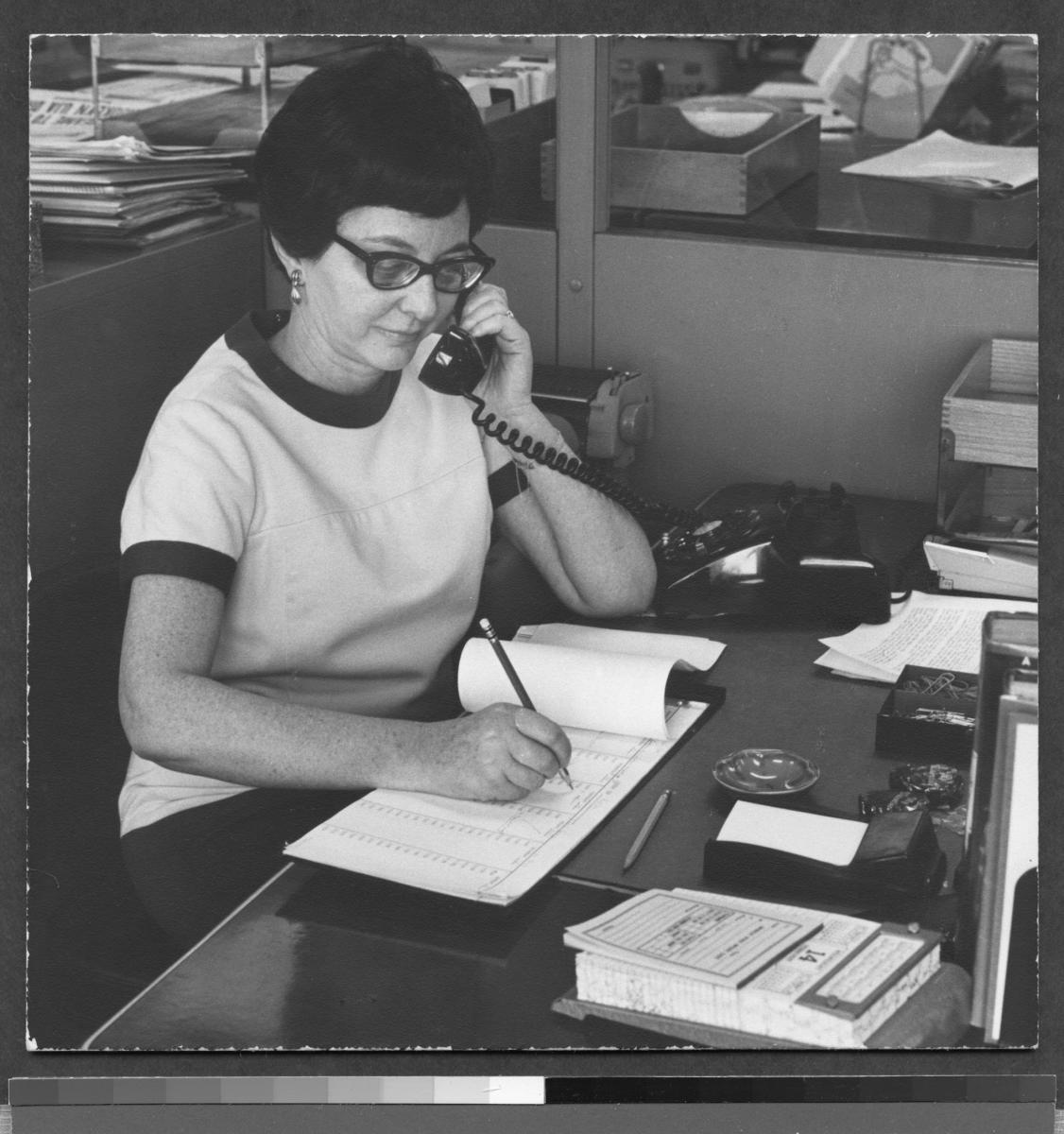 Black and white photograph of a woman writing notes while answering the phone.