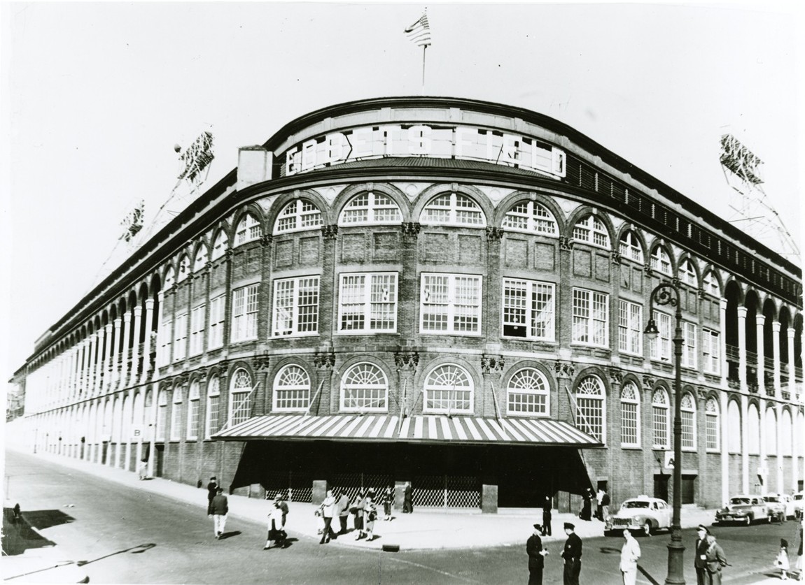 Black and white photograph of the exterior of a baseball stadium from the main entrance at the corner with several people walking in front.