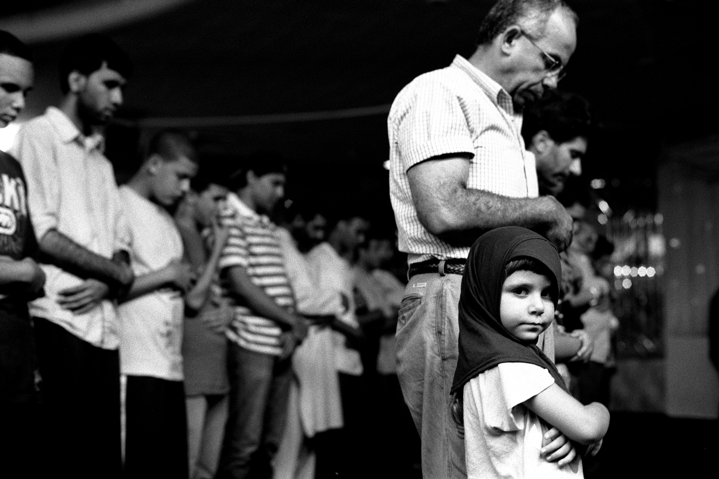 Black and white photograph of a group of men bowing their heads in prayer with a young girl wearing a headscarf in the foreground looking directly at the viewer.