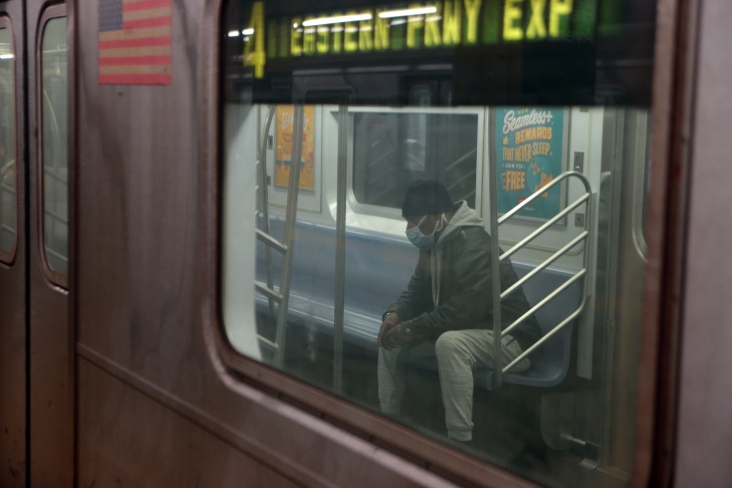Color photograph of a man wearing a surgical mask sitting inside a 4 line subway train, photographed through the subway window from the platform.