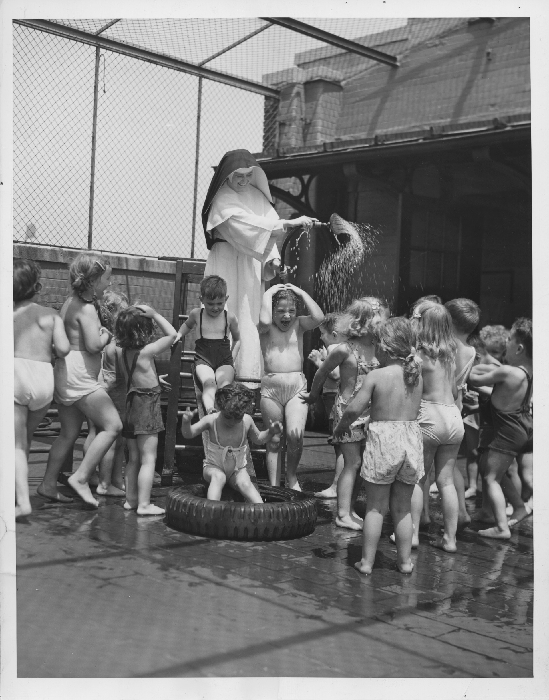 Group of young children playing on a roof, being sprinkled with water by a nun.