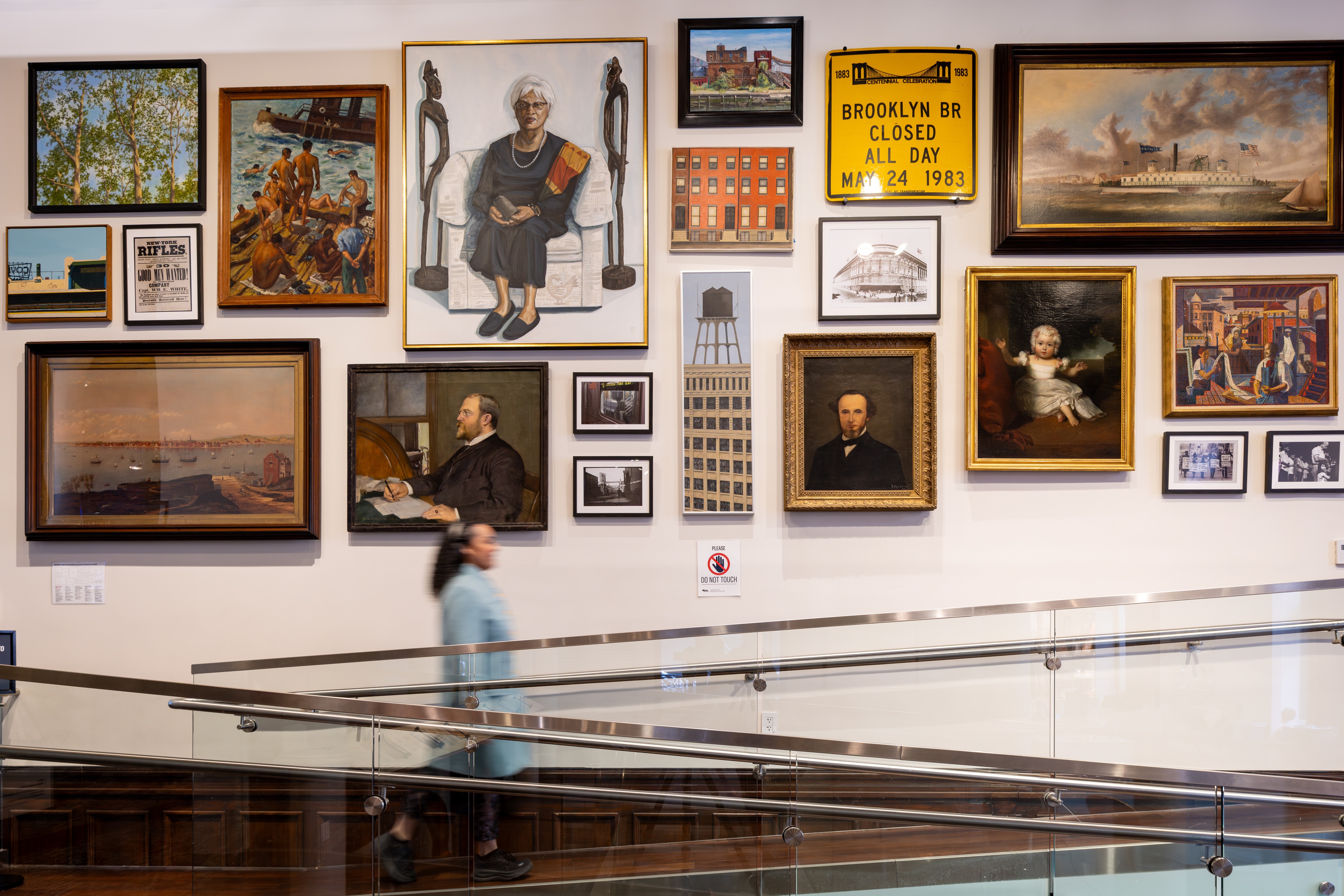 Photograph of a woman walking past a large wall with 20 artworks hung in double rows