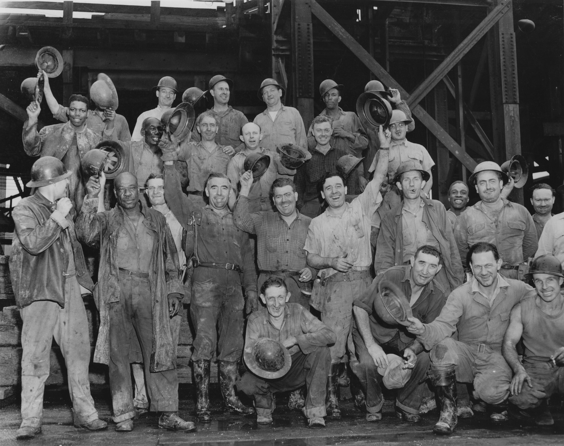 Black and white photograph of group of construction workers smiling and some waving their helmets