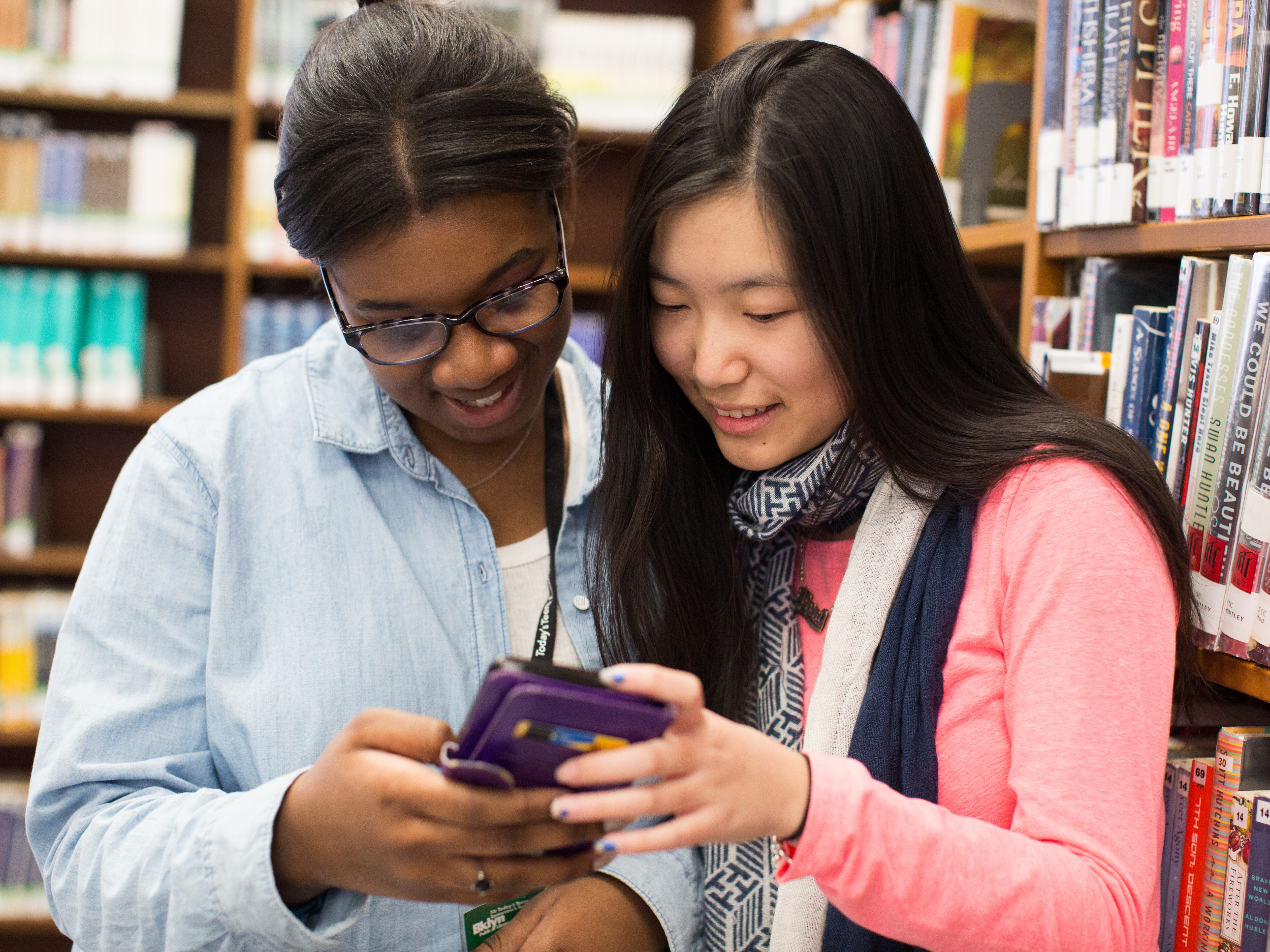 Two teen girls both smile while looking down at a phone. They are surrounded by books at a library. 
