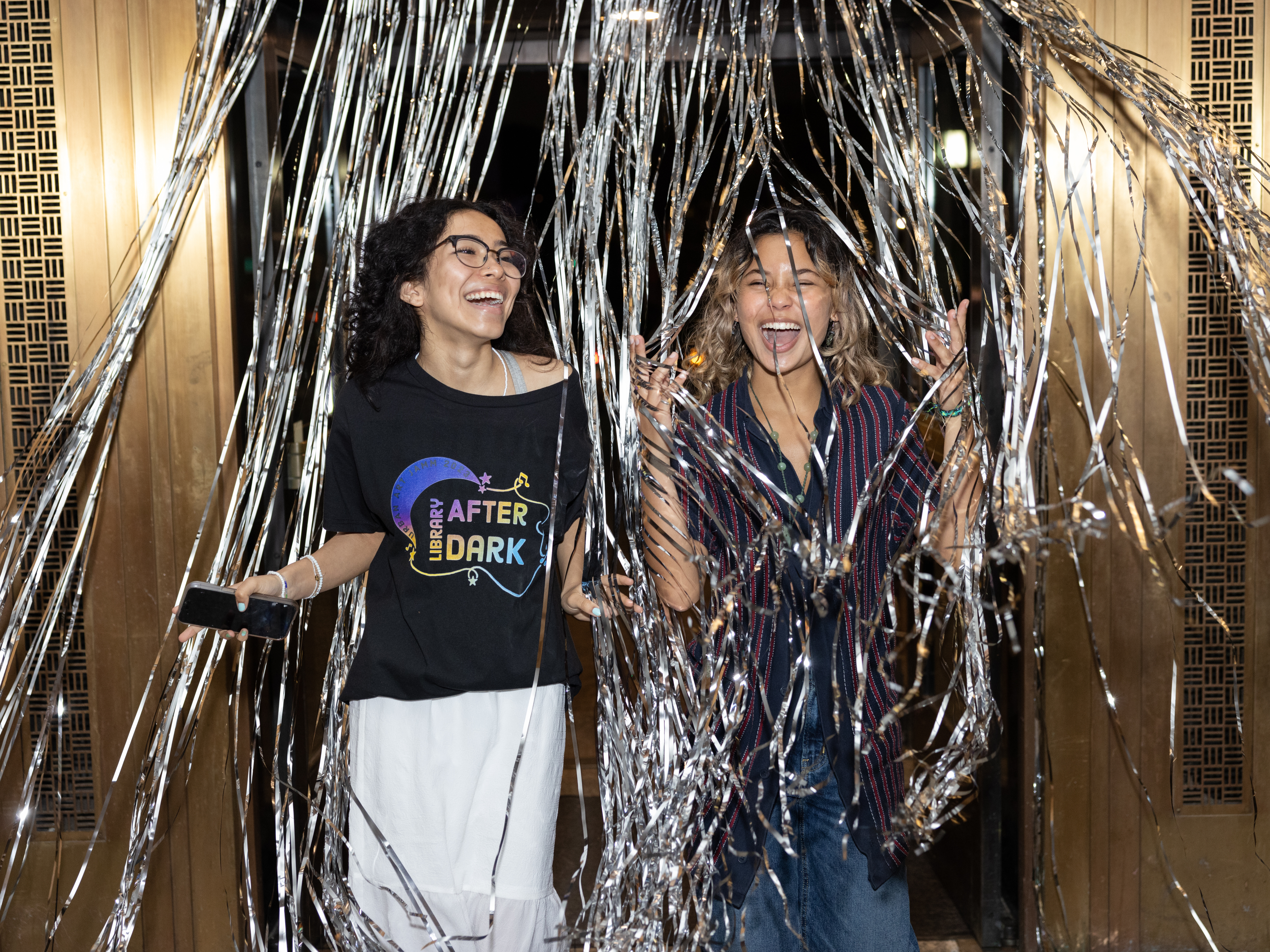 Two teen girls run out between streamers hanging over a door. They are both laughing with their hands held high. 