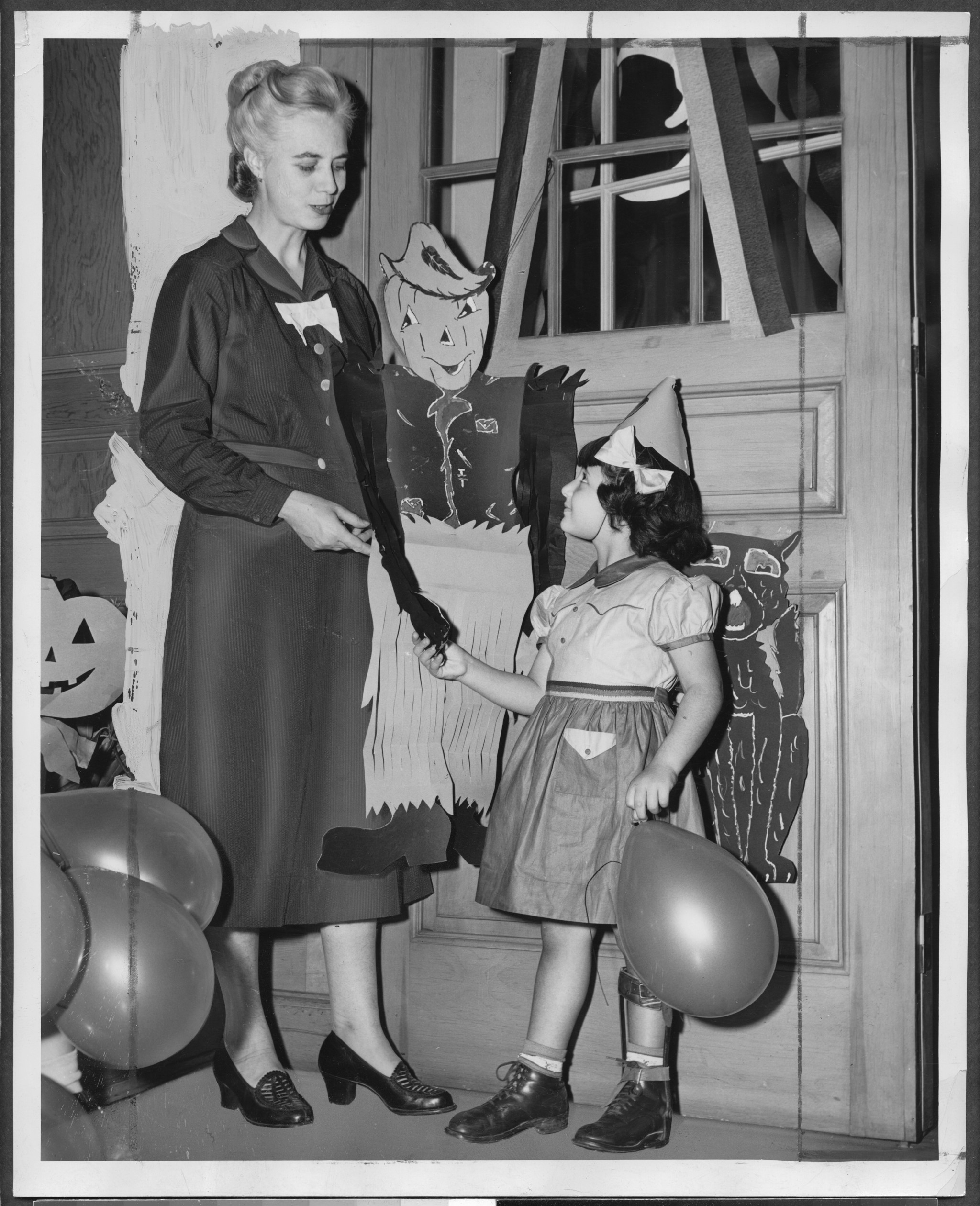 Tall woman offers the hand of a scarecrow cutout doll to a little girl in a party hat.