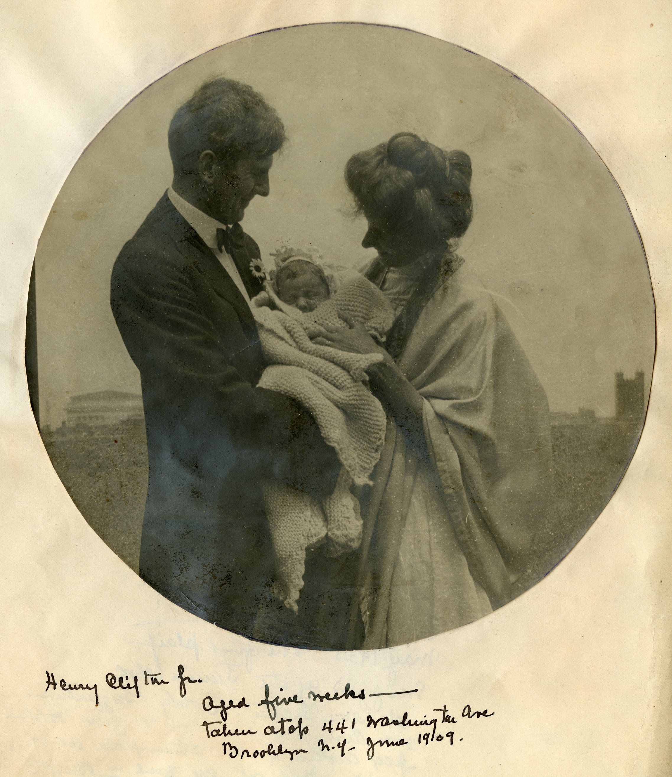 Black and white photograph of man and woman holding baby wrapped in blankets on rooftop with cityscape behind