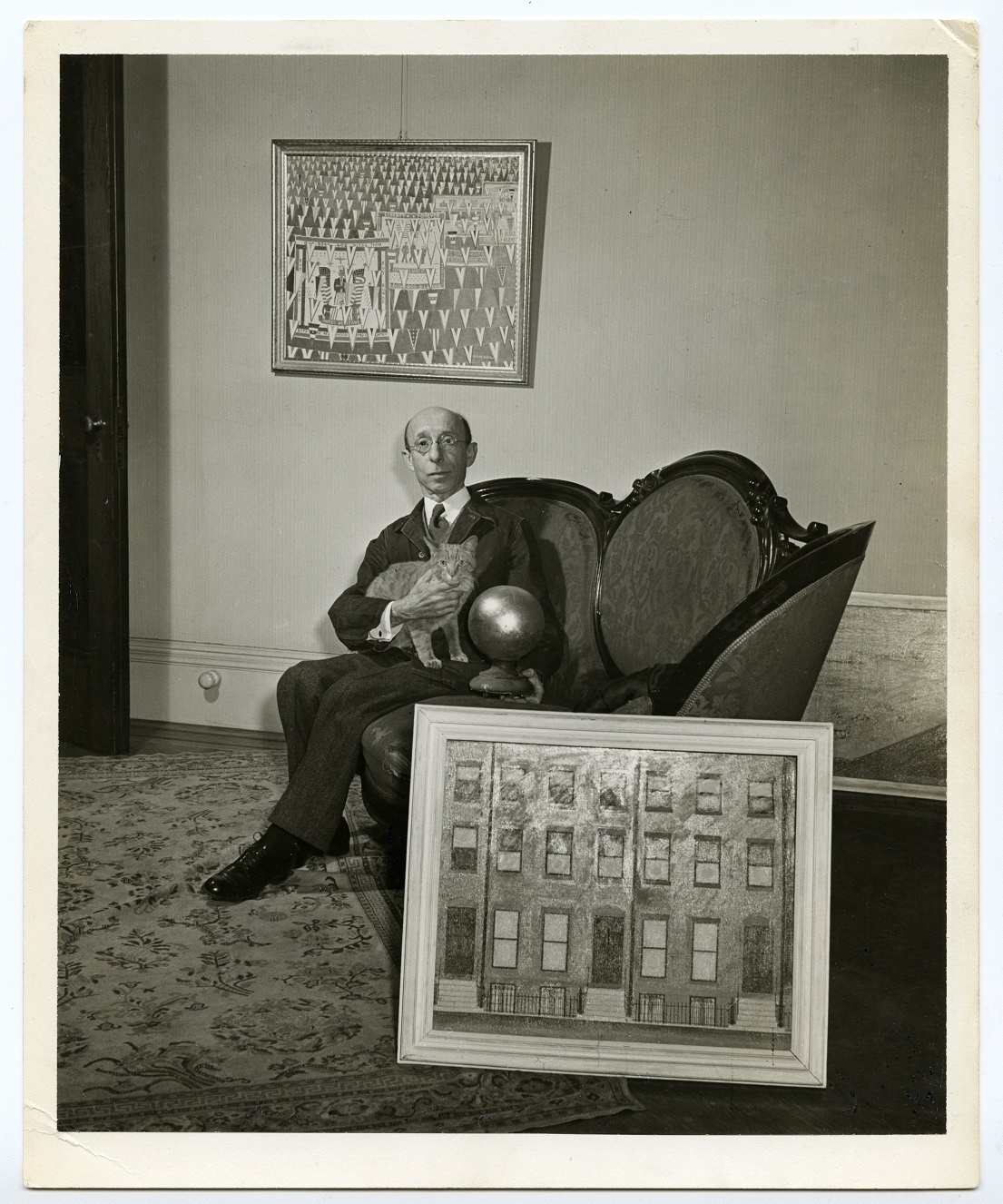 Black and white photograph of a man on a couch holding a cat with a framed painting at his feet and on the wall behind him.