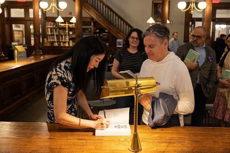 Ilyon Woo signs a man's book as she leans over a desk with a lamp in Center for Brooklyn History