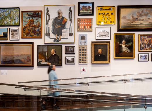 Photograph of a woman walking past a large wall with 20 artworks hung in double rows