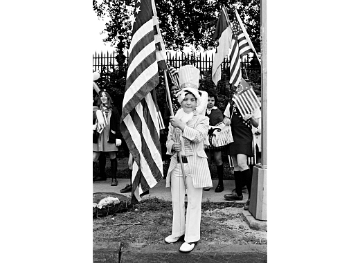A black-and-white image of a boy in a top hat and tails holding a large American flag