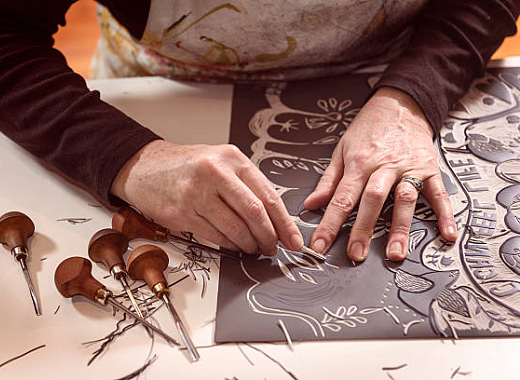 A white person uses linocut tools to carve an intricate pattern on a grey board.