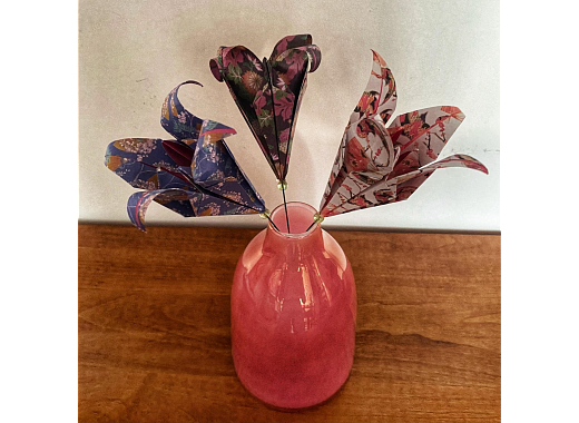 Photo of three origami lilies on wire stems in a pink vase. The vase is sitting on a wooden table and the flowers are against a white wall.
