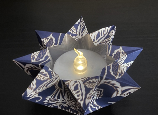 Photo of an origami votive holder with a votive candle inside (tea light size). Origami paper has a white floral design against dark blue and the photo is against a dark background.